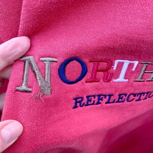 May include: Pink sweatshirt with embroidered text "NORTH" in brown, blue, red, and white, and "REFLECTION" in blue.