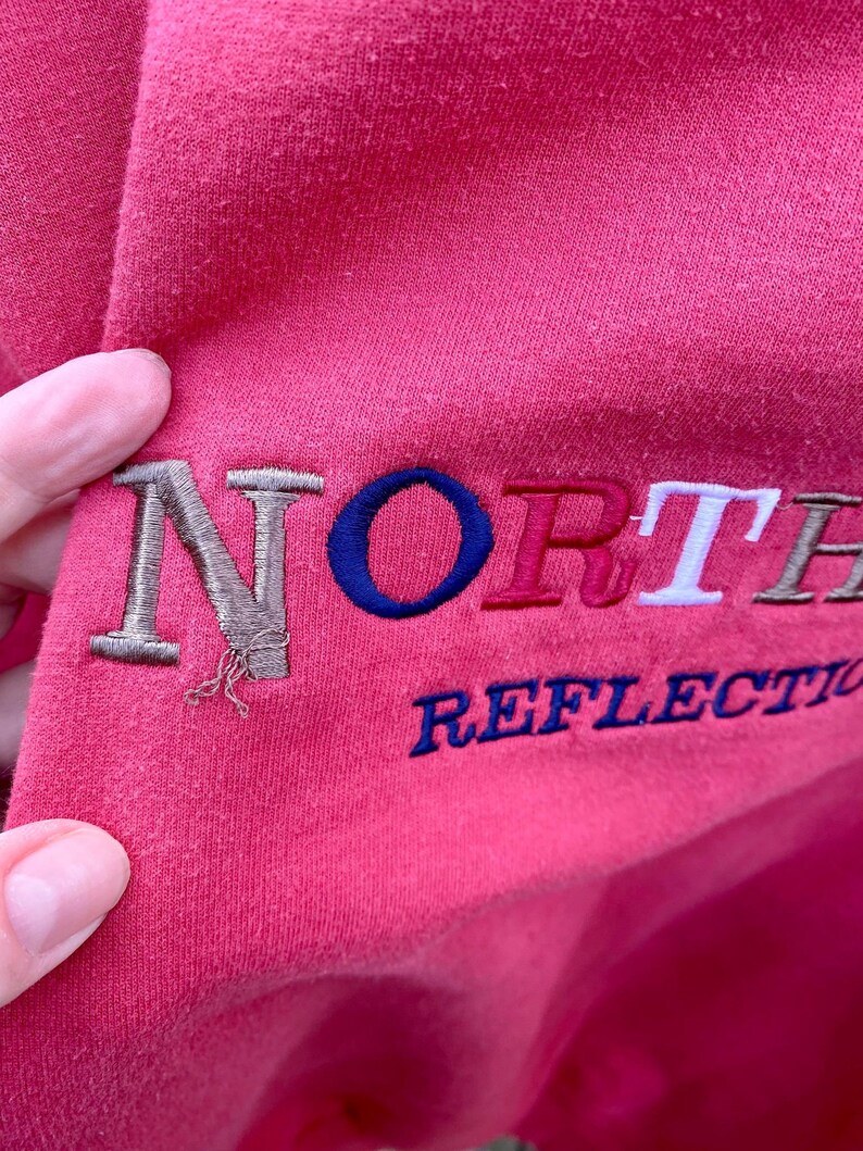 May include: Pink sweatshirt with embroidered text "NORTH" in brown, blue, red, and white, and "REFLECTION" in blue.