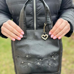 May include: Black leather handbag with embossed floral details and a heart-shaped charm. The bag features a textured handle and a structured design. A small, decorative silver heart hangs from the handle.