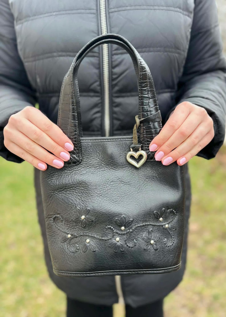May include: Black leather handbag with embossed floral details and a heart-shaped charm. The bag features a textured handle and a structured design. A small, decorative silver heart hangs from the handle.
