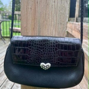 May include: Black crossbody bag with a textured flap and a silver heart-shaped clasp. The bag has a braided strap and is made of leather. The bag is displayed outdoors against a wooden post.
