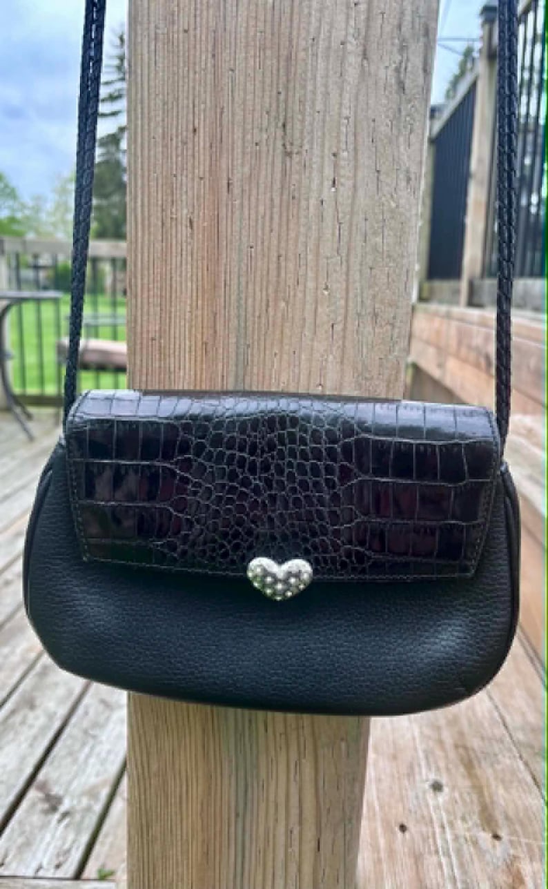 May include: Black crossbody bag with a textured flap and a silver heart-shaped clasp. The bag has a braided strap and is made of leather. The bag is displayed outdoors against a wooden post.