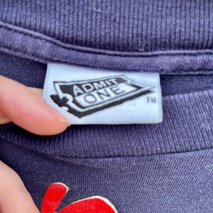 Op de afbeelding: Een marineblauw T-shirt met een rode en gele afbeelding van een cartoonfiguur. Het shirt heeft een wit label met de tekst "ADMIT ONE" en een handelsmerk.