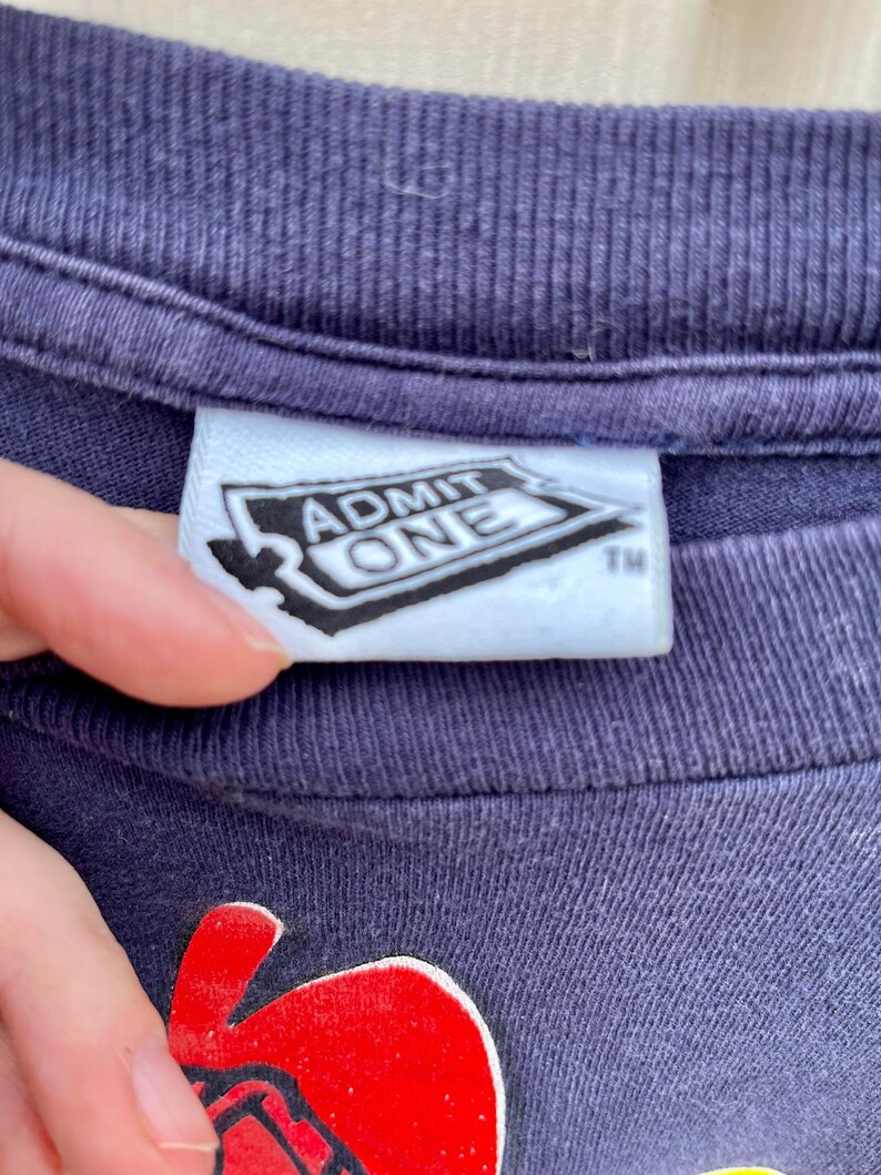 Op de afbeelding: Een marineblauw T-shirt met een rode en gele afbeelding van een cartoonfiguur. Het shirt heeft een wit label met de tekst "ADMIT ONE" en een handelsmerk.