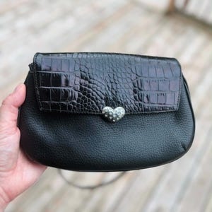 May include: Black leather clutch with a textured crocodile-patterned flap. The flap is secured with a silver heart-shaped clasp. The bag has a simple, elegant design, suitable for evening wear or special occasions.
