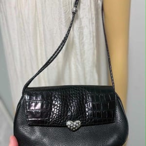 May include: Black leather crossbody bag with a textured flap and a silver heart-shaped clasp. The bag features a braided strap and a small silver bead. The bag is held by a person.