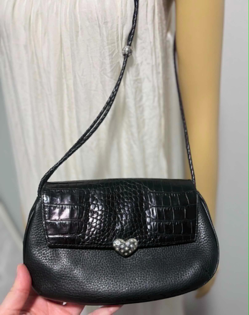 May include: Black leather crossbody bag with a textured flap and a silver heart-shaped clasp. The bag features a braided strap and a small silver bead. The bag is held by a person.