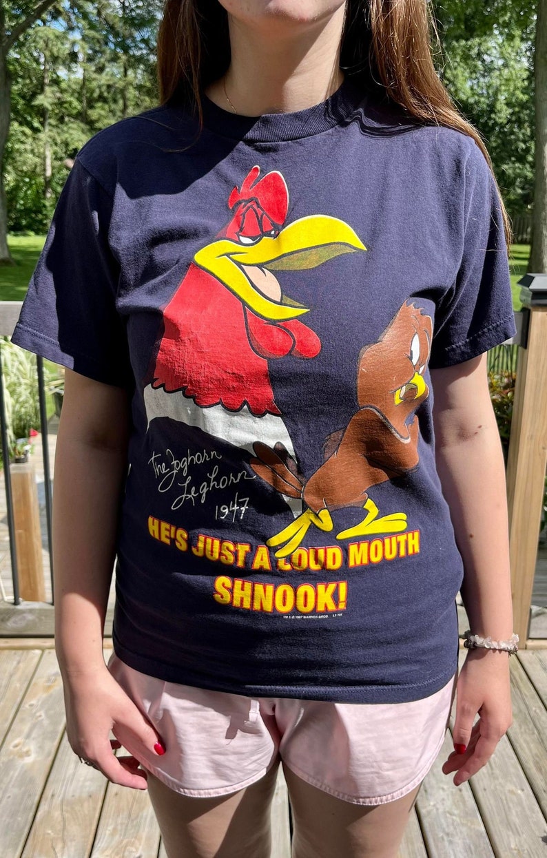 Op de afbeelding: Een marineblauw t-shirt met een cartoon-haan en een bruine vogel. Het shirt heeft de tekst "The Foghorn Leghorn 1947" en "He's just a loud mouth shnook!" erop gedrukt.