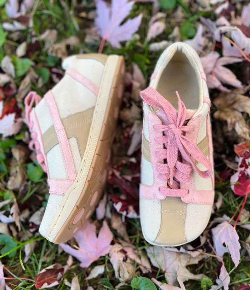 K&ouml;nnte beinhalten: Ein Paar wei&szlig;e Canvas-Sneaker mit rosa Akzenten und Schn&uuml;rsenkeln. Die Schuhe liegen auf einem Bett aus Herbstlaub.