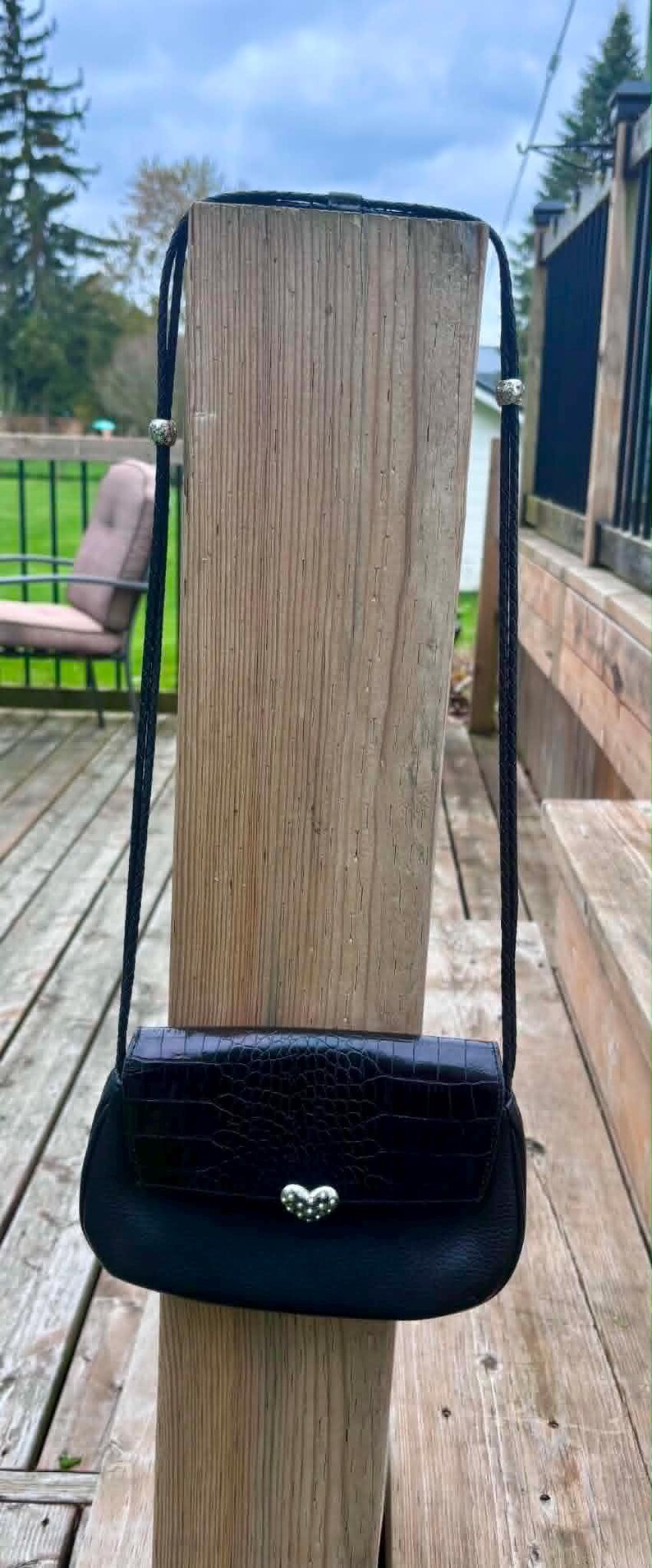 May include: Black crossbody bag with a textured, crocodile-like pattern. The bag features a silver heart-shaped clasp and a black rope strap with silver accents. The bag is hanging on a wooden post.
