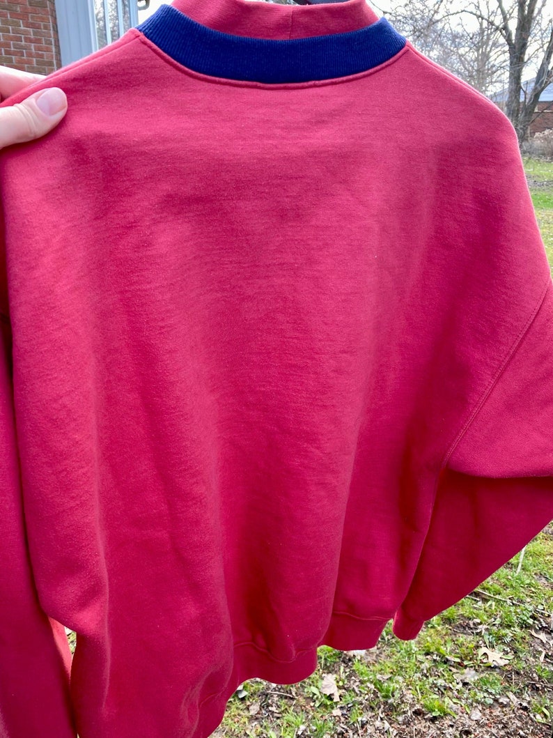 May include: A red sweatshirt with a navy blue ribbed collar. The sweatshirt is made of a soft, comfortable material.
