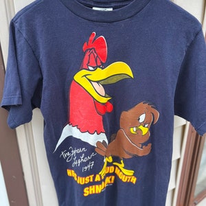 Op de afbeelding: Een marineblauw T-shirt met een cartoon-haan en een bruine vogel. De haan is rood en geel en heeft een boze uitdrukking op zijn gezicht. De vogel is bruin en heeft een verraste uitdrukking op zijn gezicht. Het shirt heeft de tekst "The Zephyr Hightower 1947 He's Just A Big Mouth SHN!" erop gedrukt.
