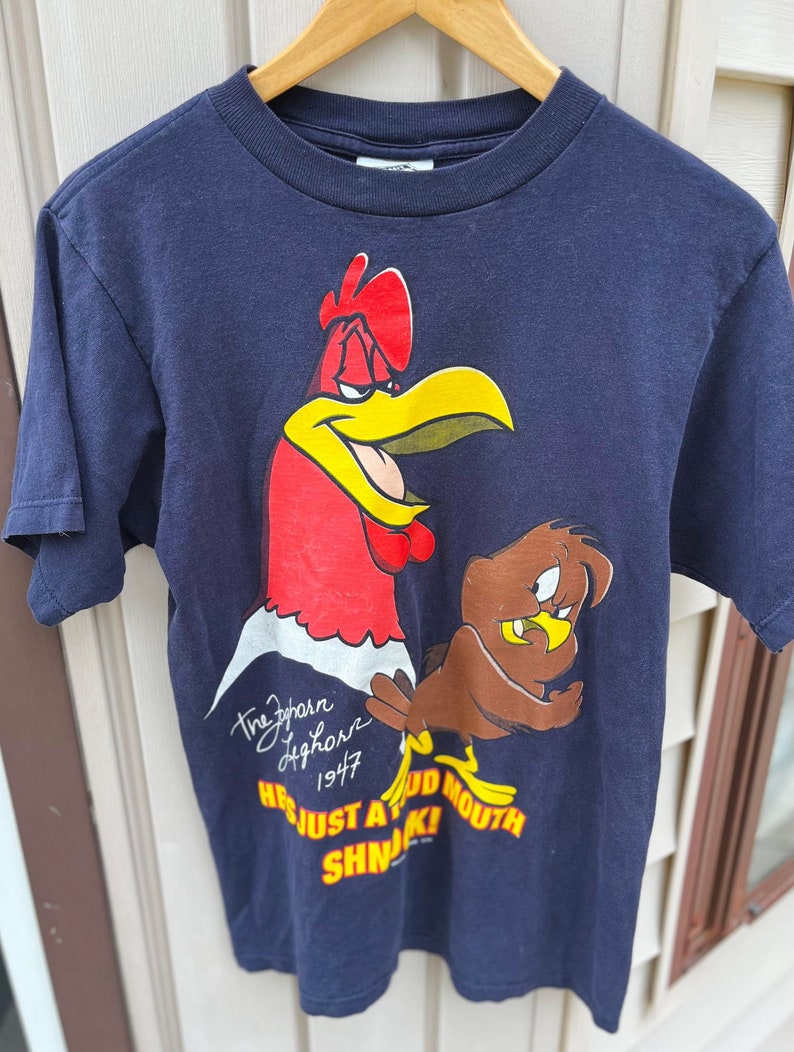Op de afbeelding: Een marineblauw T-shirt met een cartoon-haan en een bruine vogel. De haan is rood en geel en heeft een boze uitdrukking op zijn gezicht. De vogel is bruin en heeft een verraste uitdrukking op zijn gezicht. Het shirt heeft de tekst "The Zephyr Hightower 1947 He's Just A Big Mouth SHN!" erop gedrukt.