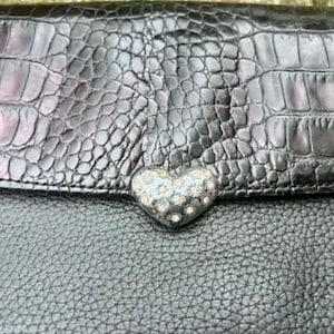 May include: Black leather handbag with a textured crocodile-like flap. The flap features a silver heart-shaped clasp adorned with small, sparkling embellishments. The bag's body has a pebbled texture.