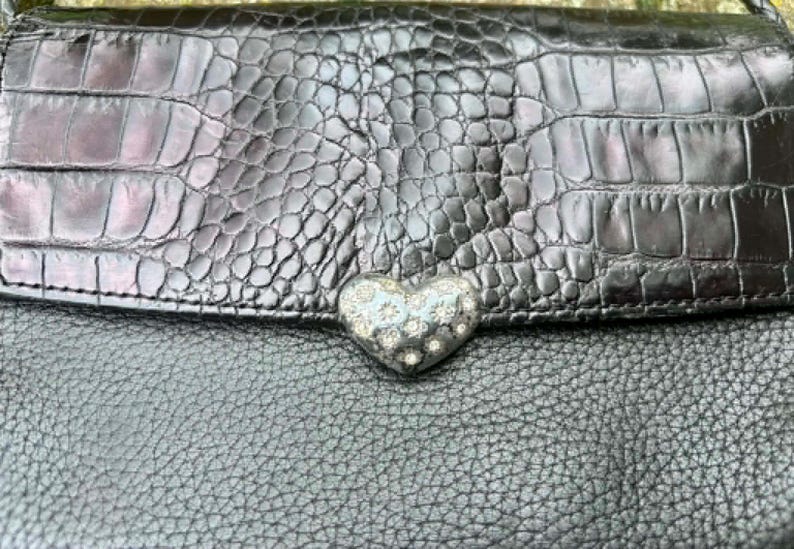 May include: Black leather handbag with a textured crocodile-like flap. The flap features a silver heart-shaped clasp adorned with small, sparkling embellishments. The bag's body has a pebbled texture.