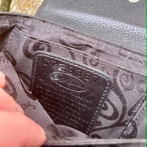 May include: Close-up of the interior of a black leather Brighton handbag. The interior features a patterned fabric lining and a small black leather tag with the number "072938" and the Brighton logo.