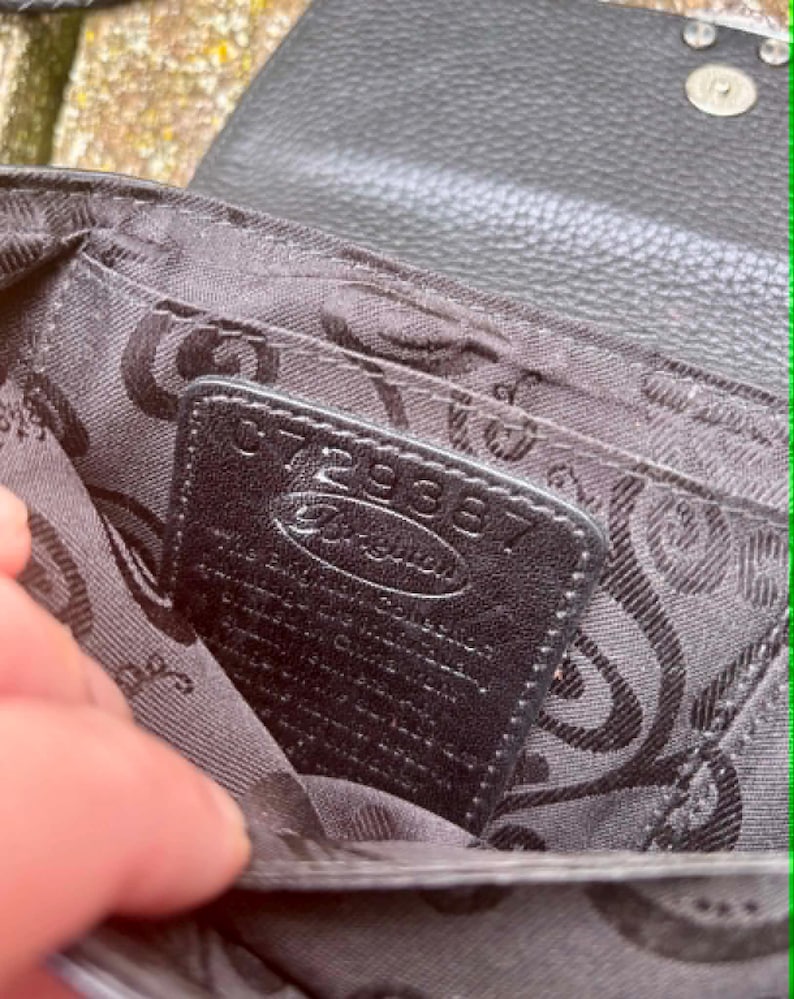 May include: Close-up of the interior of a black leather Brighton handbag. The interior features a patterned fabric lining and a small black leather tag with the number "072938" and the Brighton logo.