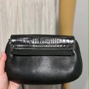 May include: A black leather clutch with a textured finish. The clutch features a flap closure with a crocodile-embossed design and a zippered pocket on the back. The bag is held by a person.