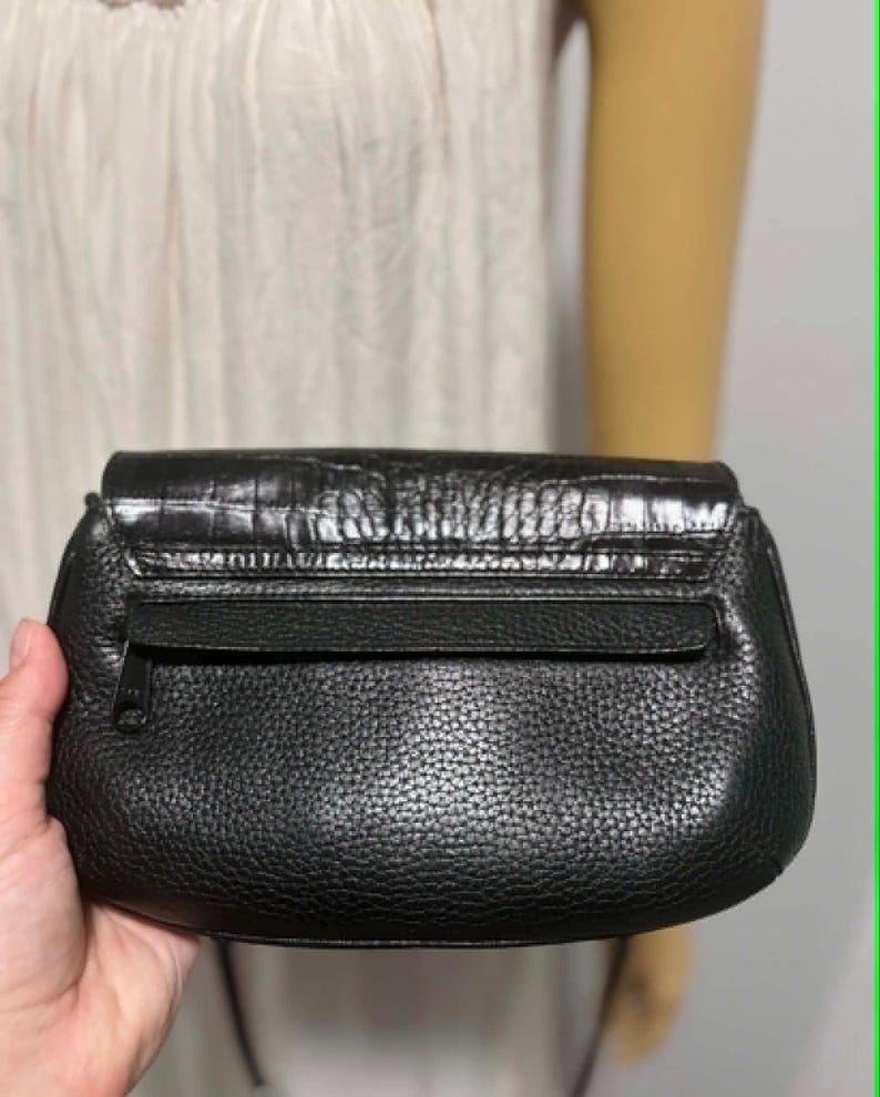May include: A black leather clutch with a textured finish. The clutch features a flap closure with a crocodile-embossed design and a zippered pocket on the back. The bag is held by a person.