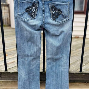 Puede incluir: Un par de pantalones vaqueros azules con bolsillos traseros adornados. Los bolsillos tienen un dise&ntilde;o floral negro con pedrer&iacute;a.