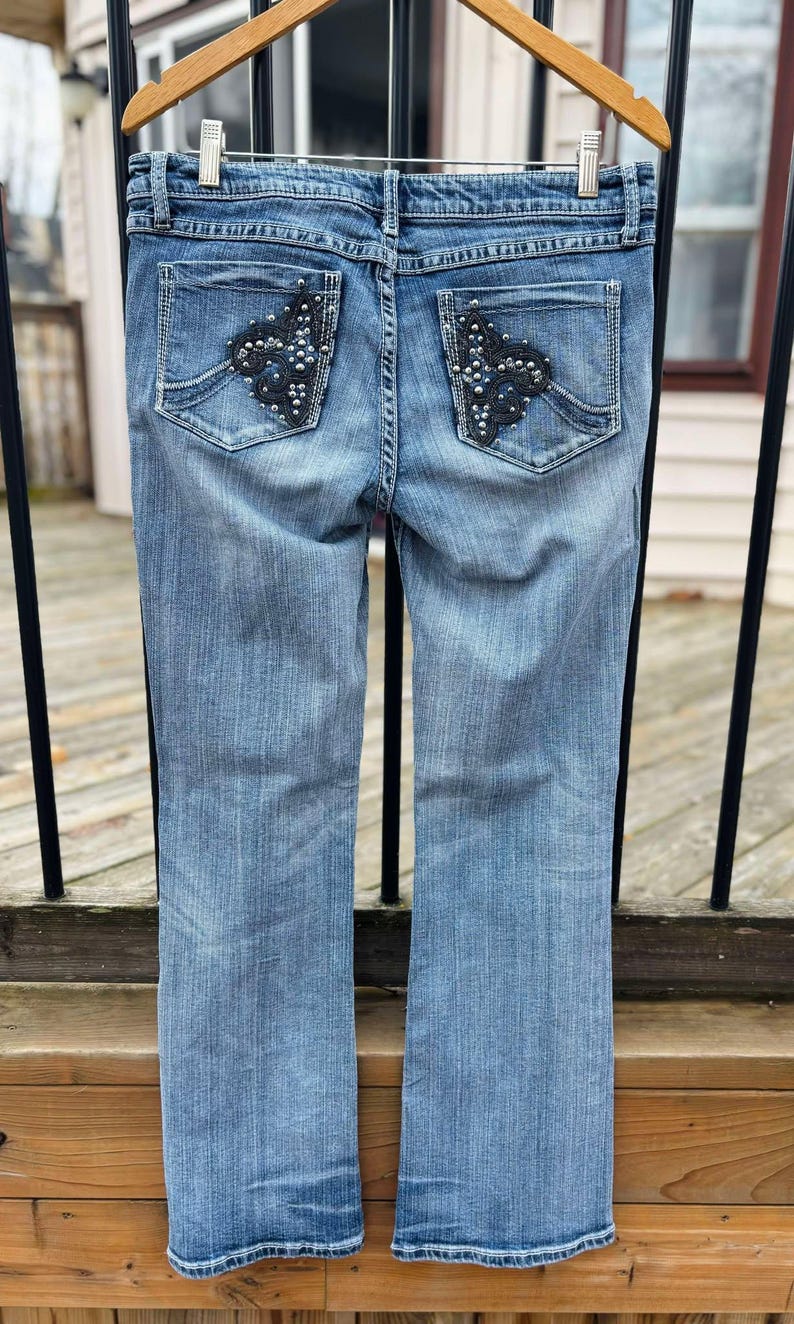 Puede incluir: Un par de pantalones vaqueros azules con bolsillos traseros adornados. Los bolsillos tienen un dise&ntilde;o floral negro con pedrer&iacute;a.