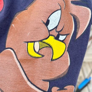Op de afbeelding: Close-up van een bruine cartoonvogel met een gele snavel en een ondeugende uitdrukking. De vogel is gedrukt op een donkerblauw T-shirt.