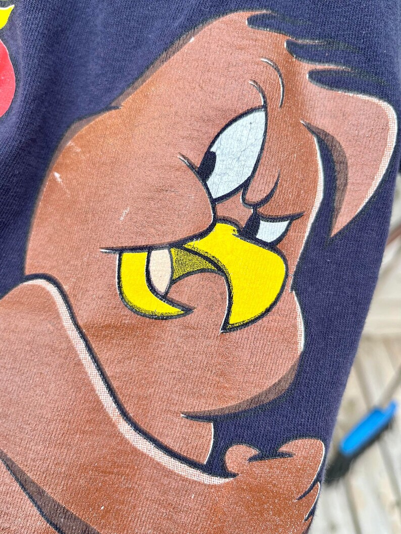 Op de afbeelding: Close-up van een bruine cartoonvogel met een gele snavel en een ondeugende uitdrukking. De vogel is gedrukt op een donkerblauw T-shirt.
