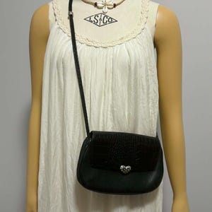 May include: Black crossbody bag with a textured flap and silver heart-shaped clasp. The bag has a braided strap and is worn over a white dress. The mannequin is wearing a necklace with a flower pendant.