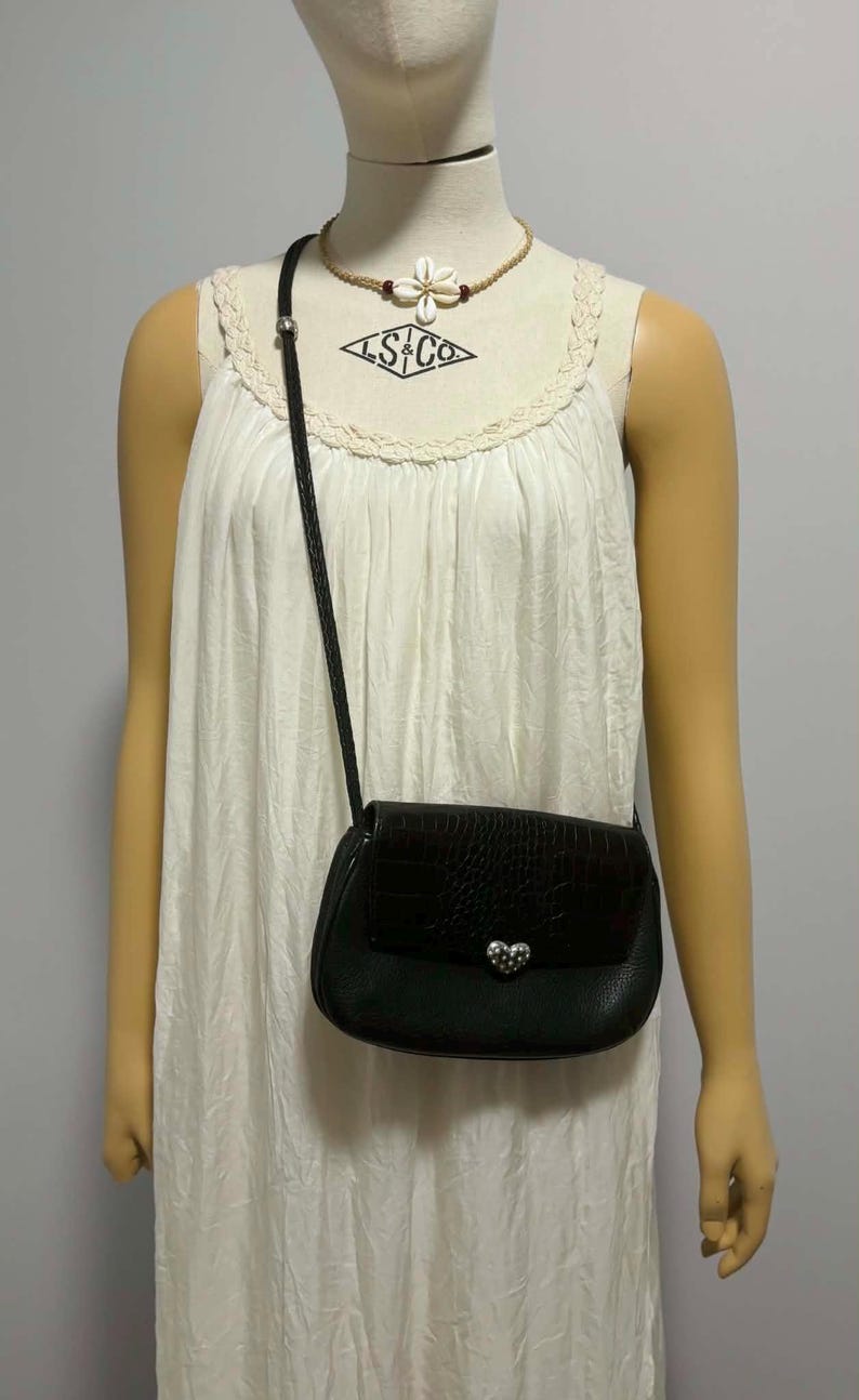 May include: Black crossbody bag with a textured flap and silver heart-shaped clasp. The bag has a braided strap and is worn over a white dress. The mannequin is wearing a necklace with a flower pendant.