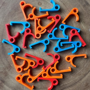 Könnte beinhalten: Eine Ansammlung kleiner, farbiger Kunststoffclips in Rot, Orange und Blau. Die Clips haben eine gebogene Hakenform mit einem kreisförmigen Loch. Sie liegen auf einer Holzoberfläche.