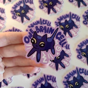 May include: A sticker with a dark blue cartoon creature with large eyes and a long tail. The text "ONE BRAIN CELL" is in a circular design on a light purple background. Other similar stickers are also visible in the image.