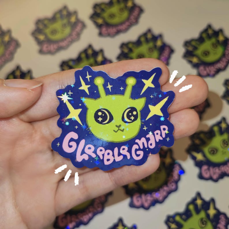 May include: A holographic sticker featuring a lime green alien with large eyes and antennae. The alien is set against a dark blue background with stars and the text "Gleebls Gnjrr" in pink and blue. Additional stickers are visible in the background.