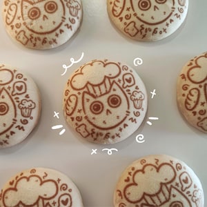 Pode incluir: Ímãs redondos de pelúcia branca com estampas marrons. Cada ímã apresenta um gato com chapéu de chef, cercado por corações, cupcakes e outros elementos decorativos. Os ímãs são dispostos em uma superfície branca.