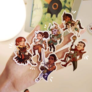 May include: A selection of seven fantasy character stickers. The stickers depict detailed illustrations of characters in various costumes, with diverse skin tones and hair colours. The stickers are held in a hand.