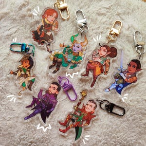 May include: A selection of acrylic character keyrings depicting fantasy figures. Each keyring has a unique design with vibrant colours and a metal clasp. The keyrings are arranged on a soft, white surface.