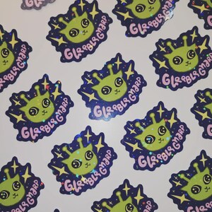 May include: A collection of stickers featuring a cartoon alien with large eyes and antennae. The alien is green with a pink mouth and is surrounded by a dark blue border with stars and the text "GLRBLS GMARP" in pink.