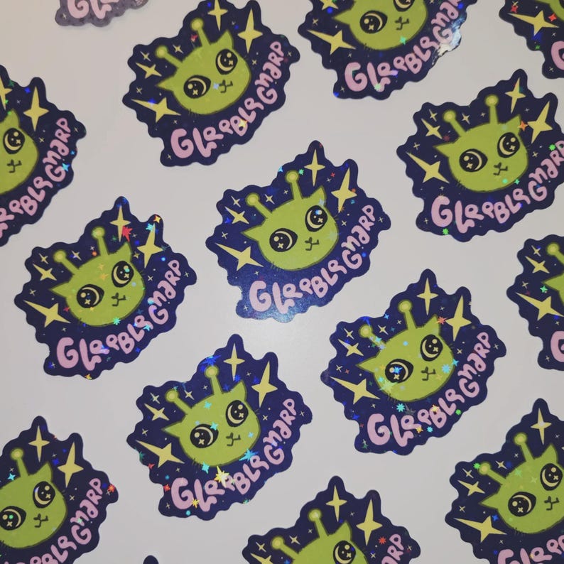 May include: A collection of stickers featuring a cartoon alien with large eyes and antennae. The alien is green with a pink mouth and is surrounded by a dark blue border with stars and the text "GLRBLS GMARP" in pink.