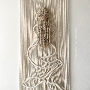 May include: Cream-coloured macrame wall hanging depicting a woman's silhouette. The design includes a headpiece, flowing hair, and a floral accent. The artwork is suspended from a wooden dowel, creating a decorative piece.