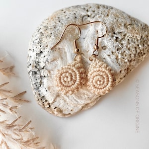 May include: A pair of beige crocheted earrings with gold hooks. The earrings are in the shape of a spiral and are hanging from a rock.
