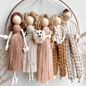 May include: A macrame wall hanging featuring five figures, including a dog, in shades of beige, brown, and white. The figures are attached to a gold hoop.