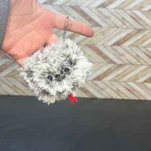 May include: A gray and white fuzzy keychain with black eyes and a red tail. The keychain is attached to a silver key ring.