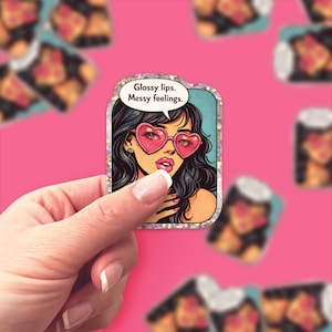 May include: A sticker featuring a pop art illustration of a woman with black hair and pink heart-shaped sunglasses. The text "Glossy lips. Messy feelings." is in a speech bubble. The sticker has a silver glitter border.