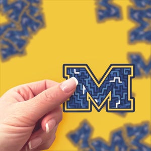 Könnte beinhalten: Ein Aufkleber in Form des Buchstabens "M" mit einem blauen Labyrinthmuster und gelben Akzenten. Der Aufkleber wird vor einem gelben Hintergrund von einer Hand gehalten, mit weiteren verschwommenen "M"-Aufklebern.