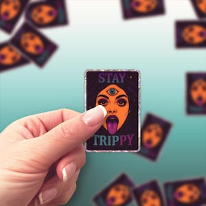 May include: A rectangular sticker with a woman's face, a third eye, and a purple tongue. The text "STAY TRIPPY" is displayed. The sticker is held by a hand against a light blue background.