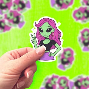 Op de afbeelding: Een sticker met een groenhuidige alien met roze haar, gekleed in een zwarte top en paarse rok, die een vredesteken geeft. De alien heeft grote zwarte ogen en gouden oorringen. De achtergrond is felgroen met andere vage alien-stickers.