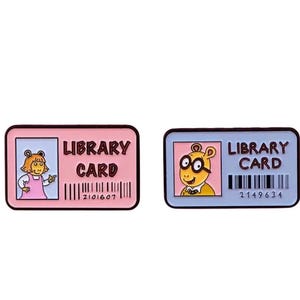 Pode incluir: Dois pinos de esmalte com personagens do programa de televisão infantil 'O BFG'. Os pinos têm a forma de cartões de biblioteca e mostram o texto 'Library Card' e um código de barras. Um pino apresenta um cartão rosa com um personagem feminino e o número de código de barras '2101607'. O outro pino apresenta um cartão azul com um personagem masculino e o número de código de barras '2149634'.