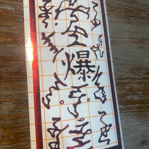 Puede incluir: Una pegatina de vinilo roja y negra con caracteres japoneses que dicen "Liberación de Fuego: Gran Técnica de Bola de Fuego". La pegatina está sobre un fondo blanco con un patrón de cuadrícula.