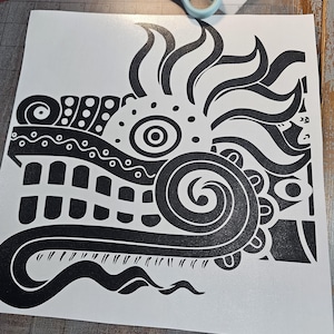 Puede incluir: Calcomanía de vinilo en blanco y negro con un diseño tribal estilizado. El diseño incluye patrones arremolinados, puntos y una espiral central, que se asemeja a una criatura mítica o una máscara. La calcomanía está sobre un fondo blanco.