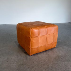 May include: A square ottoman upholstered in tan leather with a visible grid-like patchwork construction. The surface features a series of stitched leather squares creating a uniform pattern across the top and sides of this low-profile furniture piece.