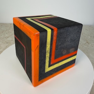 Könnte beinhalten: Ein würfelförmiges Objekt mit einer matten, schwarz texturierten Oberfläche, die mit kräftigen, geometrischen Linien in leuchtendem Orange, Gelb und Rot versehen ist. Das Design besteht aus winkligen Streifen, die sich um die Kanten legen und einen scharfen Kontrast zum dunklen, porös wirkenden Material des Hauptkörpers bilden.